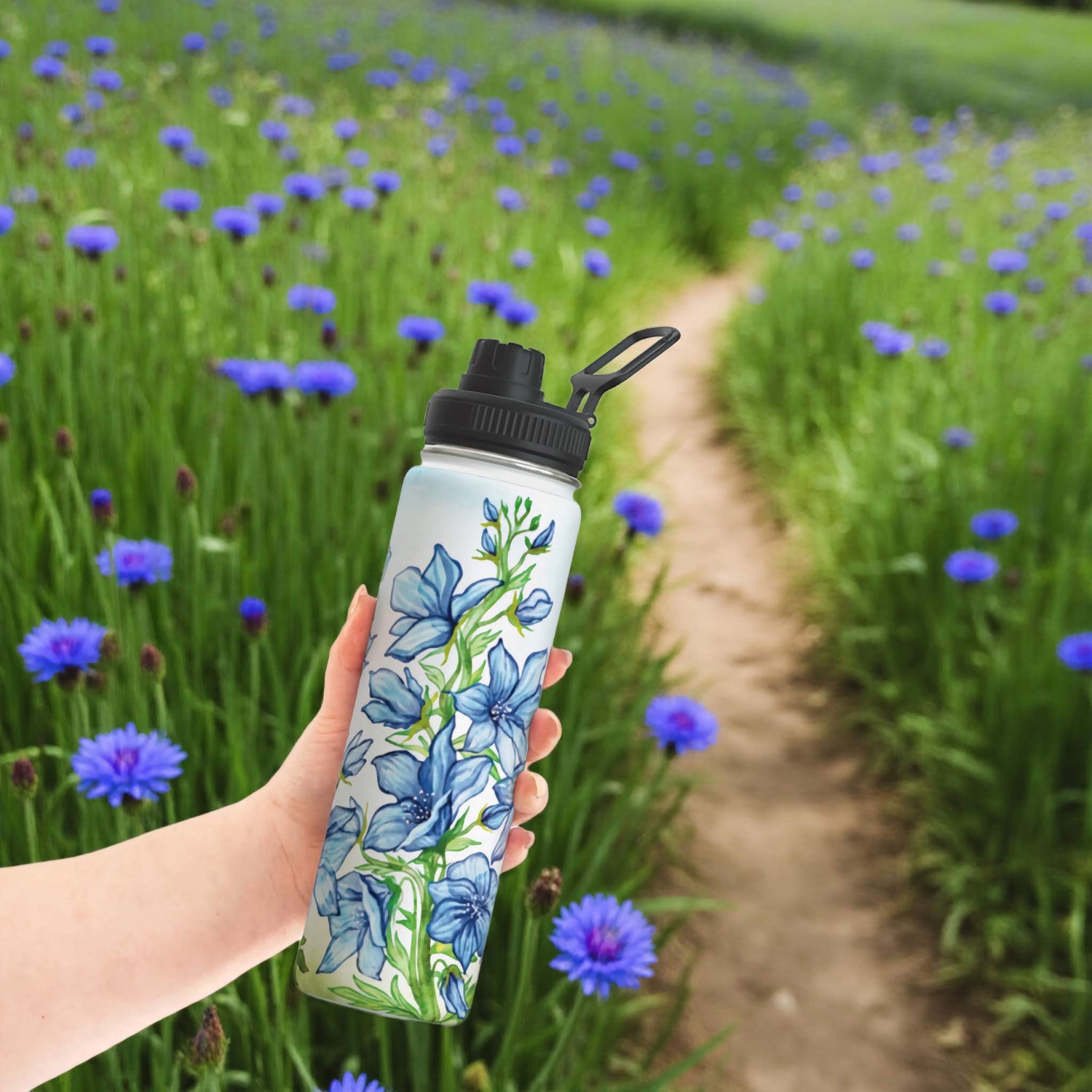 Larkspur birth flower water bottle held in hand on a sunny nature trail with blooming blue flowers and tall grass.
